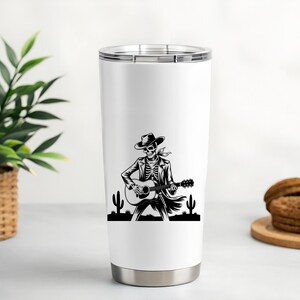 May include: White 20 oz stainless steel tumbler featuring a black and white illustration of a skeleton playing a guitar, wearing a cowboy hat and jacket. The design includes cacti silhouettes. The tumbler has a silver rim and base.