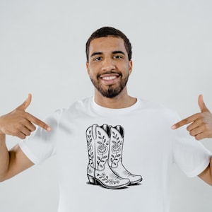 May include: White t-shirt featuring a black and white illustration of cowboy boots with floral designs. The boots have a pointed toe and a stacked heel. The design is centered on the shirt.