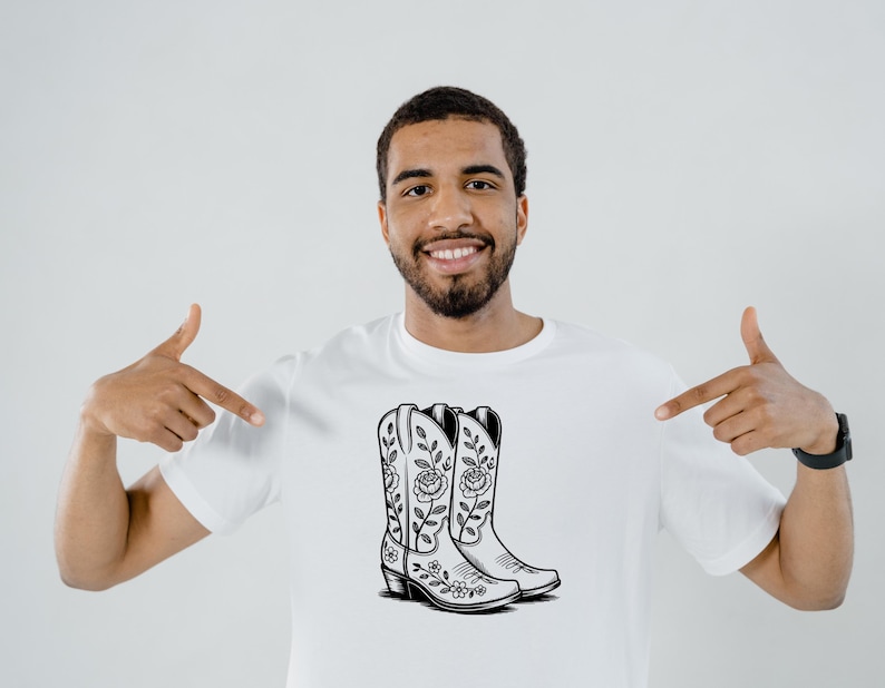 May include: White t-shirt featuring a black and white illustration of cowboy boots with floral designs. The boots have a pointed toe and a stacked heel. The design is centered on the shirt.