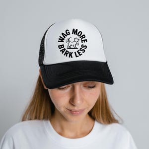 May include: A black and white trucker hat with the text "WAG MORE BARK LESS" in a circular design. The hat has a white front panel and a black brim and mesh back. The hat is worn by a person.