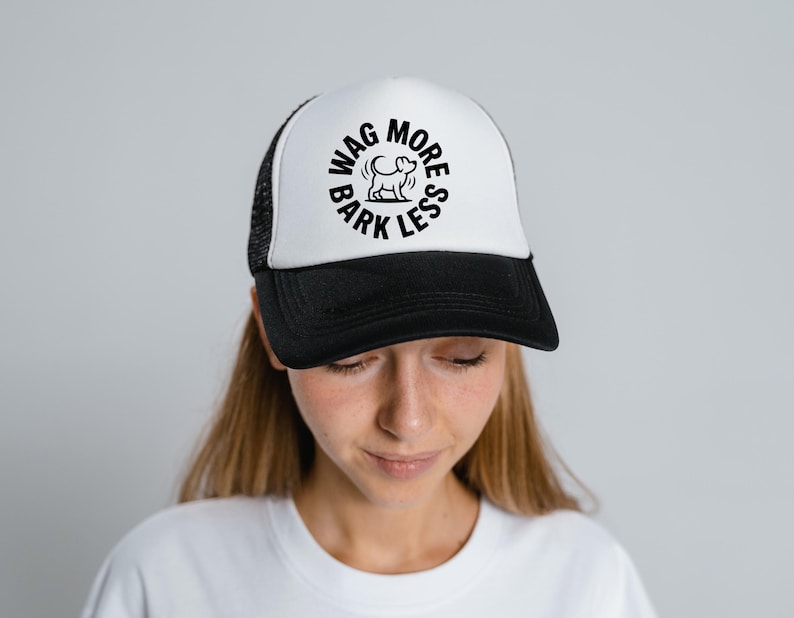 May include: A black and white trucker hat with the text "WAG MORE BARK LESS" in a circular design. The hat has a white front panel and a black brim and mesh back. The hat is worn by a person.