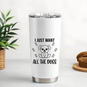 May include: A white stainless steel tumbler with a black graphic of a cartoon dog and the text "I JUST WANT ALL THE DOGS". The tumbler has a silver lid and base. A green plant in a woven basket and cookies on a wooden plate are in the background.