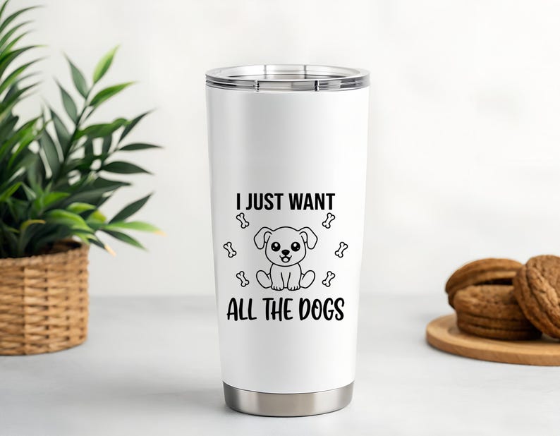 May include: A white stainless steel tumbler with a black graphic of a cartoon dog and the text "I JUST WANT ALL THE DOGS". The tumbler has a silver lid and base. A green plant in a woven basket and cookies on a wooden plate are in the background.