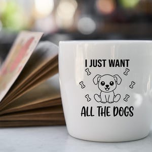 May include: A white ceramic mug with the text "I JUST WANT ALL THE DOGS" in black. The mug features a cartoon dog illustration and bone graphics. The mug is sitting next to an open book.