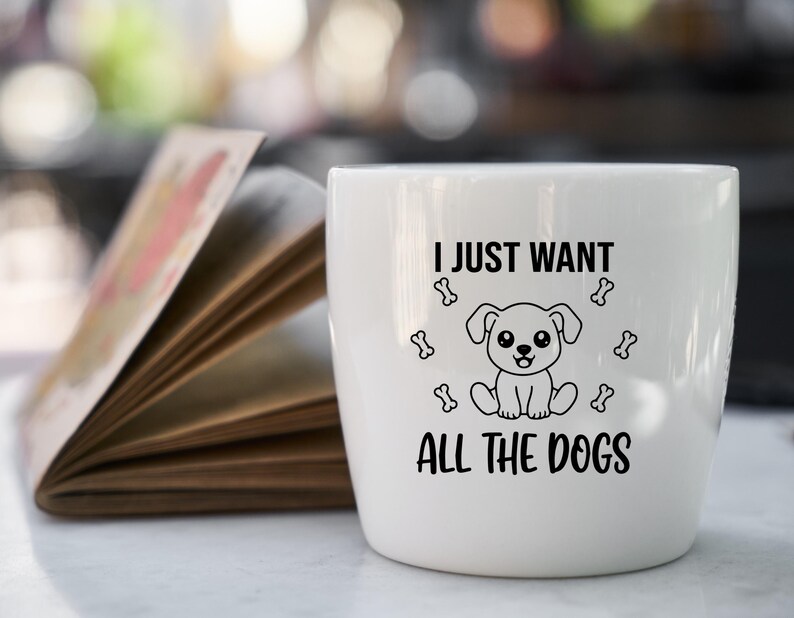 May include: A white ceramic mug with the text "I JUST WANT ALL THE DOGS" in black. The mug features a cartoon dog illustration and bone graphics. The mug is sitting next to an open book.