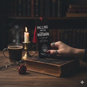 May include: A black bookmark with the text "FALLING FOR THE villain one page at a time" and a red tassel. The bookmark is held over a vintage book. A lit candle, a cup of coffee, and a rose are also on the wooden table.