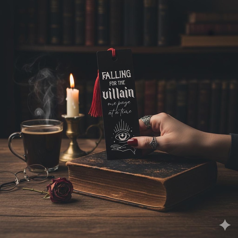 May include: A black bookmark with the text "FALLING FOR THE villain one page at a time" and a red tassel. The bookmark is held over a vintage book. A lit candle, a cup of coffee, and a rose are also on the wooden table.