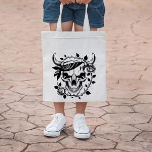 May include: White tote bag featuring a black skull design with horns, a bandana, and roses. The graphic is centered on the bag, which is held by a person wearing denim shorts and white sneakers.
