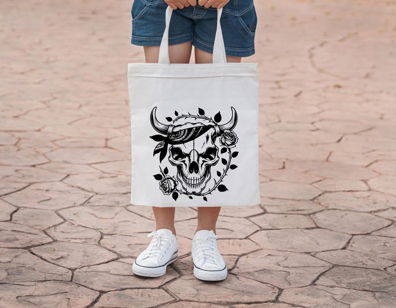 May include: White tote bag featuring a black skull design with horns, a bandana, and roses. The graphic is centered on the bag, which is held by a person wearing denim shorts and white sneakers.