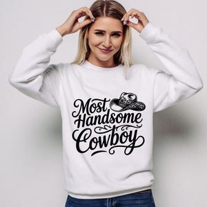 May include: White sweatshirt with the text "Most Handsome Cowboy" in a black, decorative font, along with a cowboy hat illustration. The sweatshirt is a crew neck style.