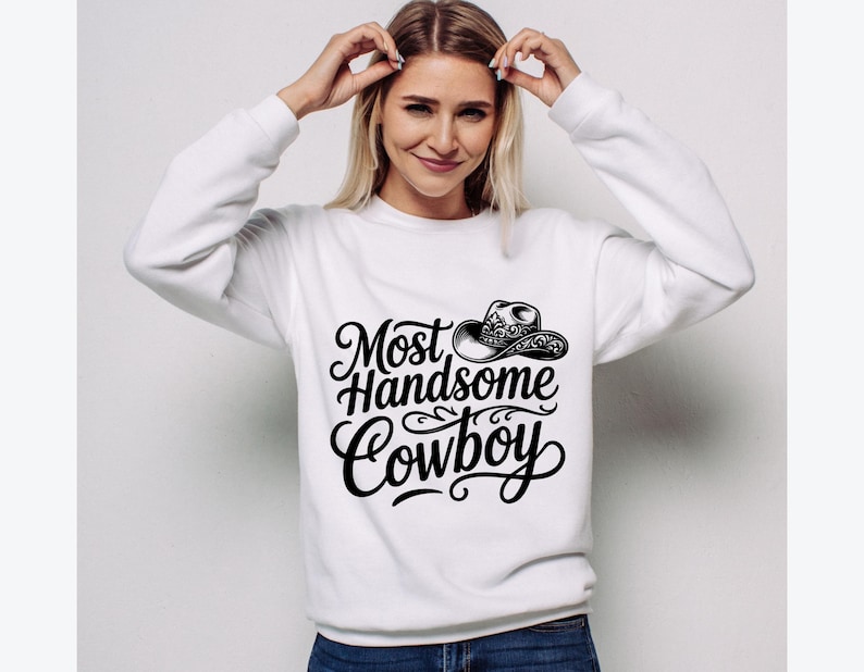 May include: White sweatshirt with the text "Most Handsome Cowboy" in a black, decorative font, along with a cowboy hat illustration. The sweatshirt is a crew neck style.