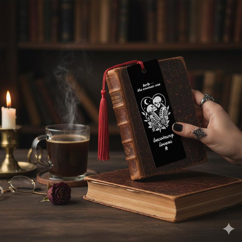 May include: A black bookmark with a red tassel is placed in a vintage book. The bookmark features a heart-shaped design with skeletons and the text "hrb... the enemies are becoming lovers *."