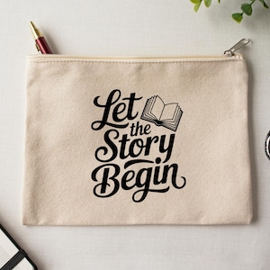 May include: A beige canvas zippered pouch with the text "Let the Story Begin" in black script and a small book illustration. A red and gold pen is in the zipper. The pouch is on a white surface with scissors and greenery.