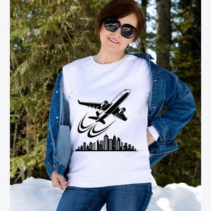 May include: White crew neck sweatshirt featuring a black graphic of an airplane taking off over a city skyline. The sweatshirt is worn with a denim jacket and blue jeans. The image is set against a backdrop of trees and snow.