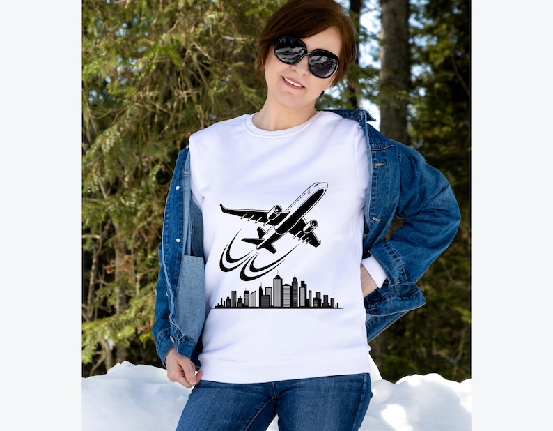 May include: White crew neck sweatshirt featuring a black graphic of an airplane taking off over a city skyline. The sweatshirt is worn with a denim jacket and blue jeans. The image is set against a backdrop of trees and snow.