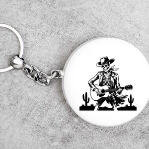 May include: A white, round keychain fob with a black and white illustration of a skeleton playing a guitar, wearing a cowboy hat and jacket. The keychain has a silver ring and chain.