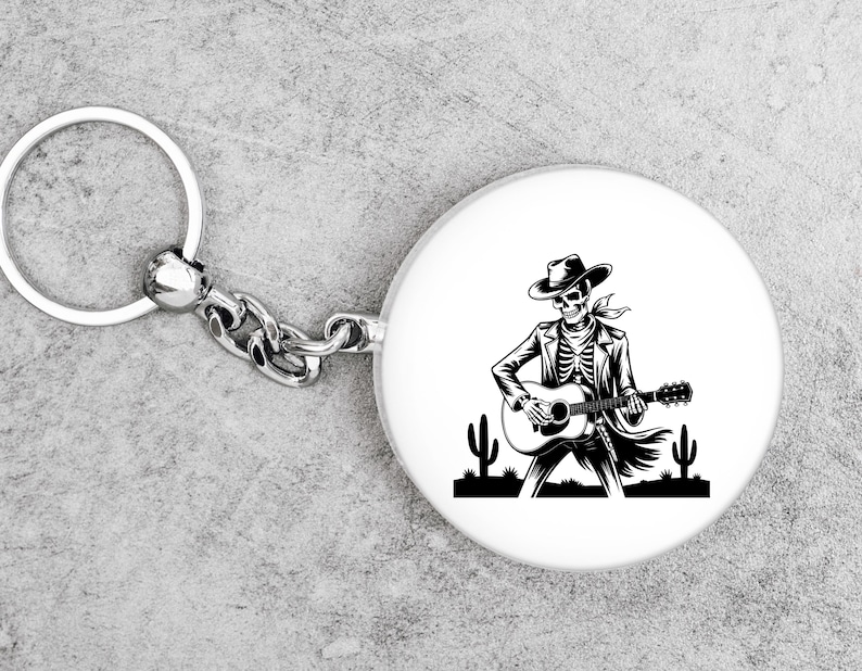 May include: A white, round keychain fob with a black and white illustration of a skeleton playing a guitar, wearing a cowboy hat and jacket. The keychain has a silver ring and chain.