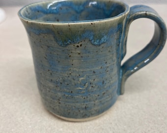 May include: A handmade ceramic mug with a blue glaze. The mug has a rounded body and a sturdy handle. The blue glaze has darker blue and green accents, giving it a unique, speckled appearance. The mug is suitable for hot or cold beverages.
