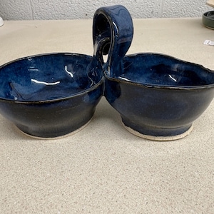 May include: A handmade ceramic double bowl server featuring a deep navy blue glaze with subtle variations in tone. The two circular bowls are connected by a central arched handle, and the base of each bowl reveals a raw, unglazed white clay rim.