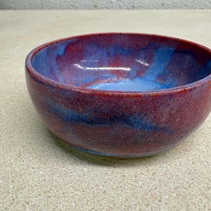 May include: A round ceramic bowl featuring a glossy glaze with a blended color palette of deep red, magenta, and vibrant blue. The interior and exterior surfaces display a smooth, mottled finish with swirling patterns of these colors.
