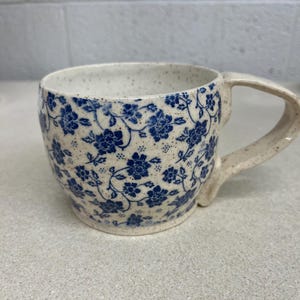 May include: A ceramic mug with a speckled beige exterior and a blue floral pattern. The mug has a curved handle and a wide opening. The floral design is a repeating pattern of small flowers and leaves.