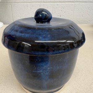 May include: A ceramic jar with a lid featuring a deep blue and black glossy glaze. The container has a rounded body and a matching lid with a small, smooth knob handle on top. The base of the jar shows a light, unglazed speckled clay texture.