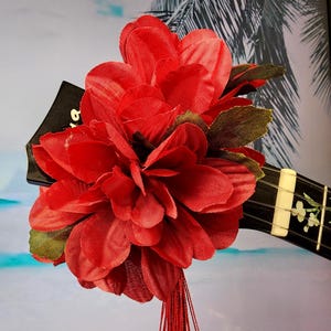 May include: A vibrant red flower arrangement with green leaves and red tassels, attached to a black ukulele. The ukulele has white frets and floral designs. The background is a blurred image of a tropical scene.