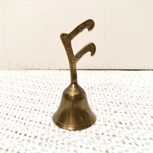 May include: A small, antique brass bell with a decorative handle. The bell has a flared base and a detailed handle with curved ends. The bell is approximately 10 cm tall and sits on a white, textured surface.