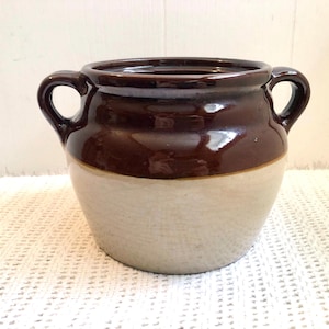 May include: A two-tone ceramic crock in brown and cream. The top half is a glossy, dark brown, and the bottom half is a matte cream colour. It features two loop handles and a wide opening. The crock has a rounded body.