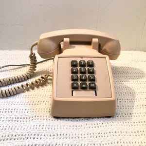 Può includere: Un telefono beige vintage con una cornetta, una base con tastierino numerico e un cavo a spirale. Il telefono ha una base rettangolare con tasti numerici neri e una culla per la cornetta.