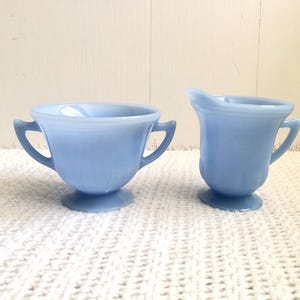 May include: A light blue vintage sugar bowl and creamer set. The sugar bowl has two handles and a pedestal base. The creamer has a handle and a spout. Both pieces are made of glass and have a ribbed design.