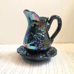 May include: A dark iridescent glass jug and matching saucer set. The jug features a handle and a grape cluster design. The set has a glossy finish with a colour-shifting effect, displaying hues of blue, purple, and green.