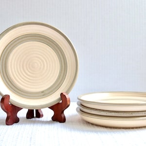 Peut inclure: Ensemble de quatre assiettes en céramique avec un fond blanc et des anneaux gris concentriques. Les assiettes sont empilées les unes sur les autres et sont exposées sur un support en bois.