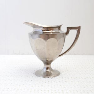 May include: A silver creamer with a fluted design and a handle. The creamer has a round base and a spout.