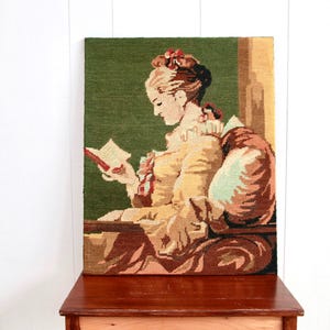 Peut inclure: Une tapisserie murale représentant une femme en robe jaune lisant un livre. La tapisserie a un fond vert et est encadrée dans un cadre en bois.