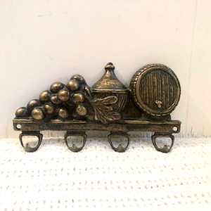 Peut inclure: Porte-clés mural de couleur bronze avec une grappe de raisin, une bouteille de vin et un tonneau. Le porte-clés comporte quatre crochets pour suspendre des clés ou d'autres petits objets. Le design rappelle une cave à vin.