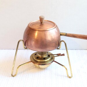 Puede incluir: Olla de fondue de cobre con asa y tapa de madera, sobre un soporte de latón. La olla tiene forma redondeada y un pequeño pomo de madera en la tapa. El soporte tiene tres patas.