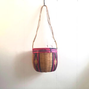 May include: A woven basket bag with a shoulder strap. The basket is tan with pink and dark purple accents. The bag is hanging against a white wall.