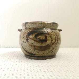 Peut inclure: Un pot en céramique fait à la main avec une palette de couleurs tachetées et terreuses. Le pot présente un motif en spirale brun foncé et deux petites poignées. La base est d'une nuance de brun plus foncée. Idéal pour la décoration intérieure.