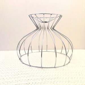 May include: A chrome wireframe decorative bowl with an hourglass shape. The bowl has a wide base and a flared top, with vertical wires creating the form. The bowl is on a white, textured surface.