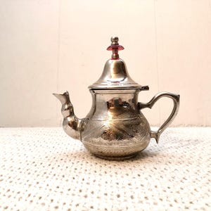 Może przedstawiać: Srebrny czajnik z misternymi wzorami kwiatowymi i czerwonym guzikiem. Czajnik ma zakrzywiony uchwyt i dzióbek o unikalnym designie. Jest to przedmiot dekoracyjny, prawdopodobnie do serwowania herbaty.