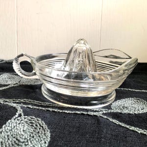 May include: A clear glass citrus juicer with a ribbed bowl, a pointed reamer, and a handle. The juicer is designed for easy citrus fruit juice extraction. The design is vintage and functional.