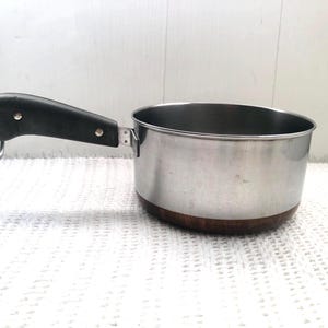 May include: A stainless steel saucepan with a copper base and a black plastic handle. The handle has a hook on the end.