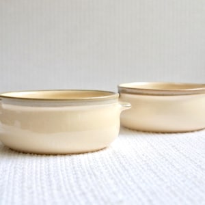 May include: Two cream-colored ceramic bowls with brown rims and handles. The bowls are on a white surface.