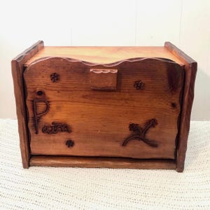 Könnte beinhalten: Brotdose aus Holz mit natürlichem Finish. Die Box hat geschnitzte Blumen- und Blattmotive sowie das Wort "POTA" auf der Vorderseite. Der Deckel hat eine wellenförmige Kante und einen kleinen Holzverschluss.