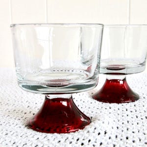 Peut inclure: Deux coupelles à dessert en verre transparent avec des pieds rouges. Les coupelles sont vides et posées sur une surface en crochet blanc.