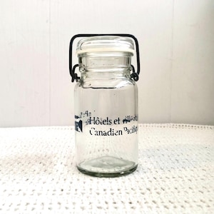 Peut inclure: Un bocal en verre transparent avec un couvercle blanc et une poignée en fil de fer noir. Le bocal porte une inscription bleue et blanche : "An Hôtels et villes Canadien Pacifique". Le bocal est posé sur une surface blanche crochetée.