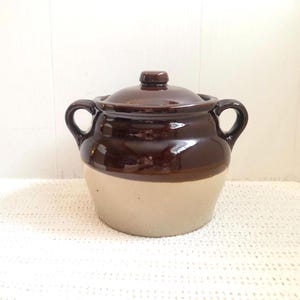May include: A two-tone ceramic cooking pot with a lid. The top half is a glossy dark brown, and the bottom half is a matte beige. It has two loop handles and a small knob on the lid. The pot is designed for cooking and serving.