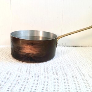 May include: A copper-colored saucepan with a long, brass-colored handle. The interior of the pan is silver, and it appears to contain liquid. The pan is set against a white background.