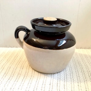 May include: A vintage stoneware crock with a dark brown top and handle, and a cream-coloured base. The crock has a matching lid with a cork stopper. The handle is curved, and the crock is likely used for food storage.